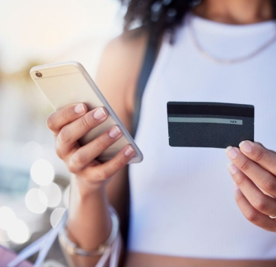 Afiliación mes de mayo 2024 Una persona sosteniendo un teléfono móvil en una mano y una tarjeta de crédito en la otra, preparándose para realizar una transacción en línea. La imagen sugiere comodidad y facilidad en las compras digitales.