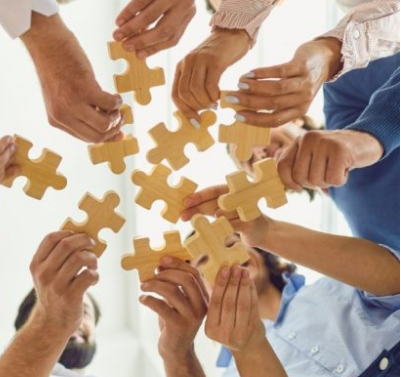 Un grupo de personas sosteniendo piezas de rompecabezas de madera, uniéndolas en un esfuerzo colaborativo. Las manos de los participantes están extendidas hacia el centro, sugiriendo trabajo en equipo y cooperación. Grupo de apoyo y psicoeducación, sábados. Mayo, 2024
