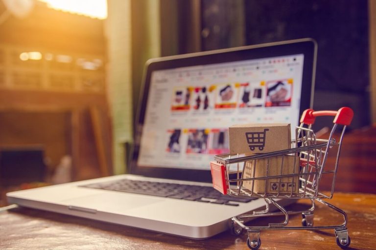 Una computadora portátil mostrando un sitio web de compras en línea con un carrito de compras pequeño lleno de paquetes colocado al lado